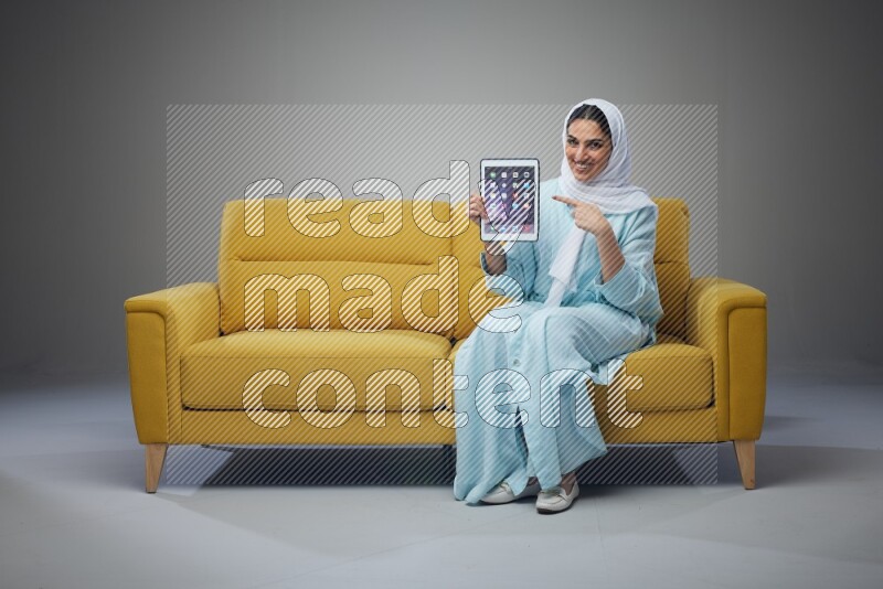 A Saudi woman wearing a light blue Abaya and white head scarf sitting on a yellow sofa and showing her tablet's screen while pointing to it eye level on a grey background