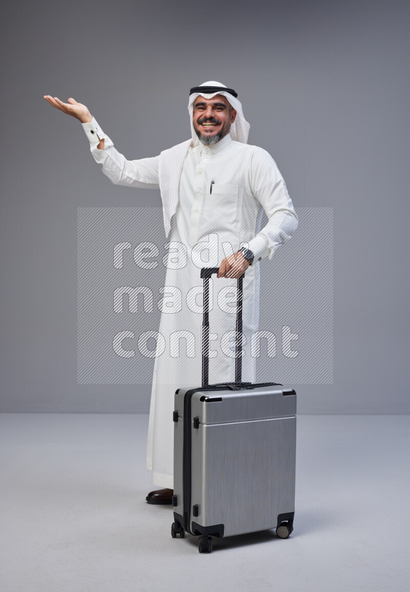 Saudi man wearing Thob and white Shomag standing holding Travel bag on Gray background