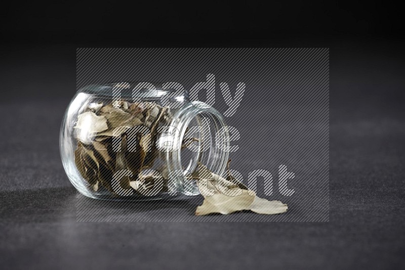 A glass spice jar full of dried bay leaves and jar is flipped and leaves spread out on black flooring