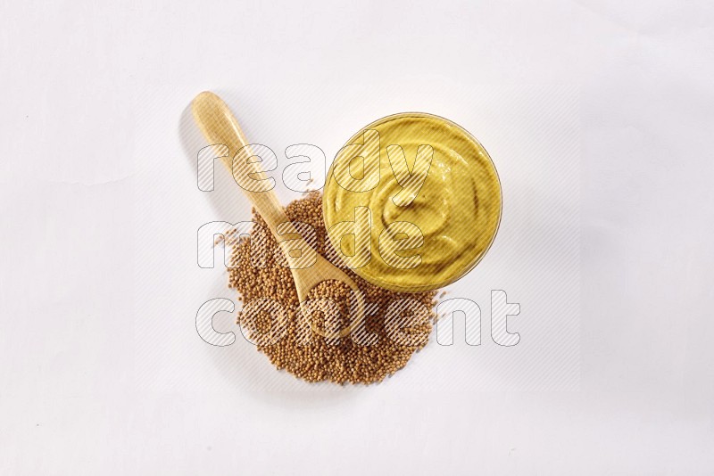 A glass bowl full of mustard paste with some of mustard seeds underneath it and a wooden spoon on white flooring