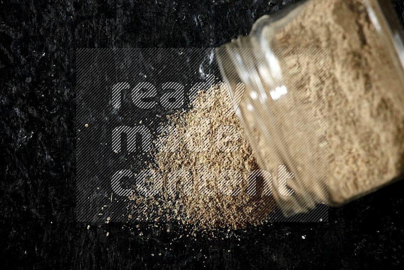 A flipped glass jar full of cardamom powder and powder spilled out of it on textured black flooring