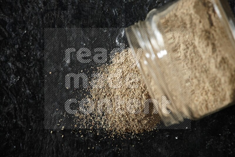 وعاء زجاجي مقلوب ممتلئ ببودرة الهيل والبودرة تناثرت منه على خلفية سوداء