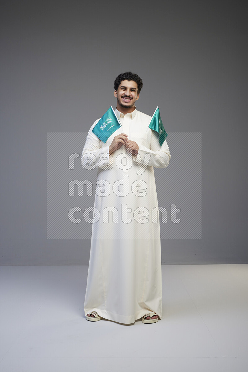 A Saudi man standing wearing thob holding small Saudi flag on gray background