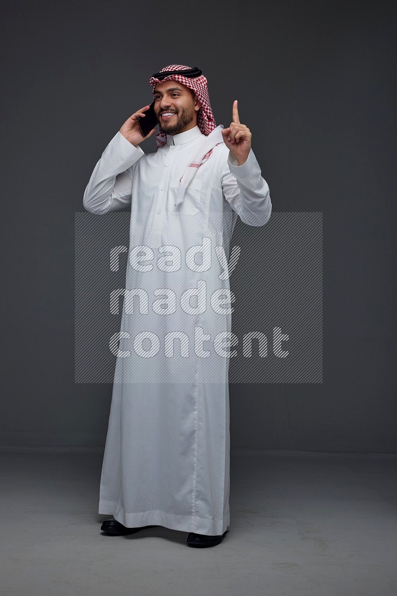 A Saudi man wearing Thobe and Shmagh talking in the phone eye level on a gray background