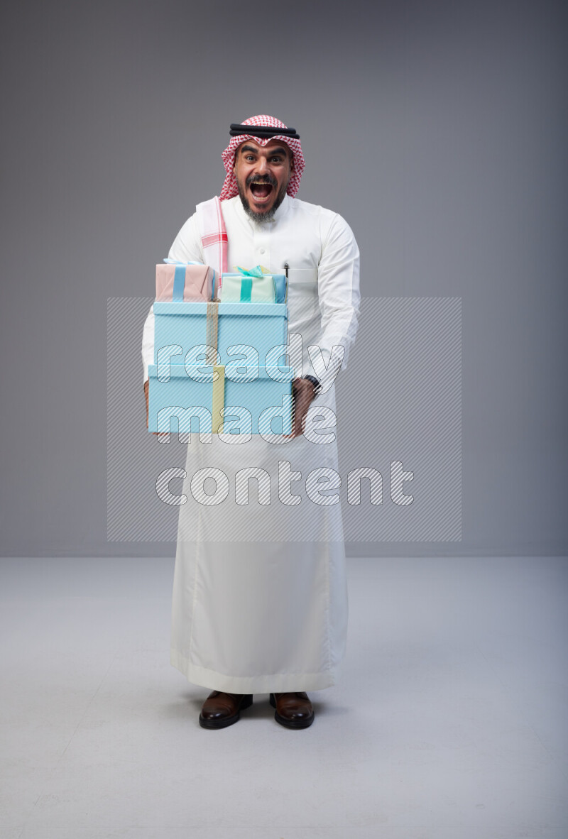 Saudi man Wearing Thob and red Shomag standing holding gift box on Gray background