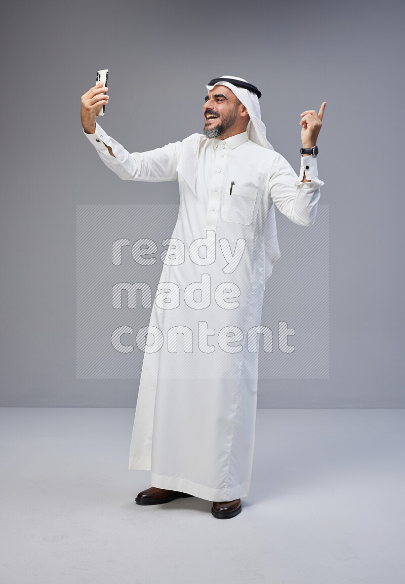 Saudi man Wearing Thob and white Shomag standing taking selfie on Gray background