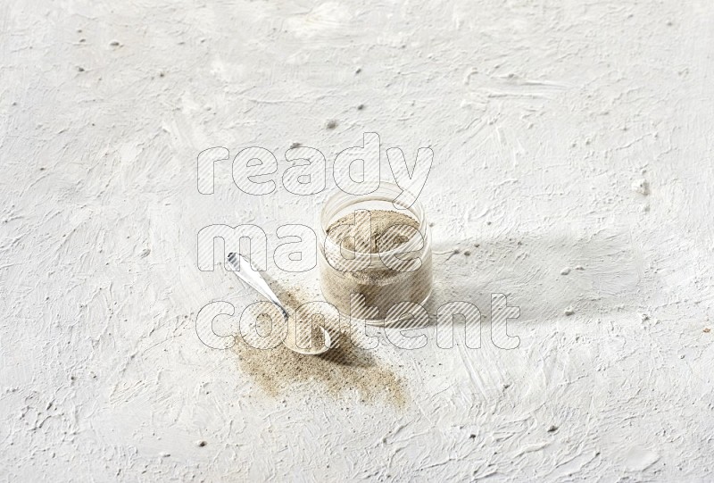 A glass jar and a metal spoon full of white pepper powder on textured white flooring