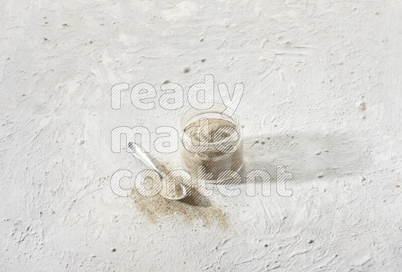 A glass jar and a metal spoon full of white pepper powder on textured white flooring