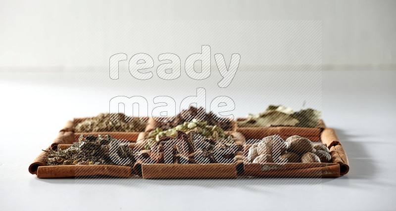 9 squares of cinnamon sticks full of cardamom in the middle surrounded by nutmeg, cinnamon, bay laurel leaves, cloves, cumin, dried ginger, dried basil and star anise on white flooring