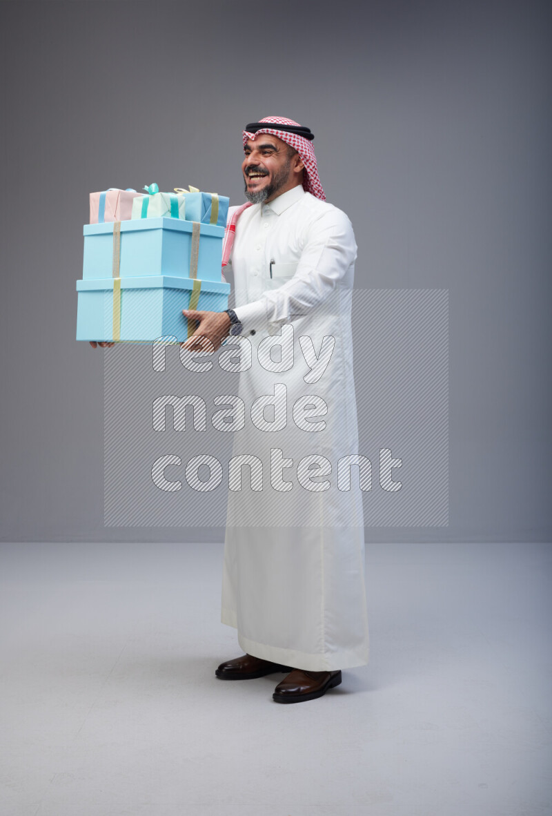 Saudi man Wearing Thob and red Shomag standing holding gift box on Gray background