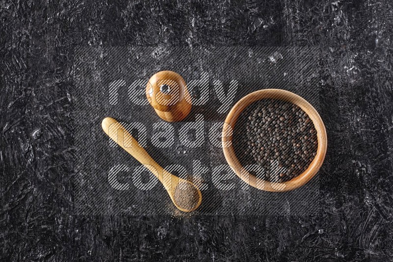 A wooden bowl full of black pepper and a wooden spoon full of black pepper powder and a wooden grinder on a textured black flooring