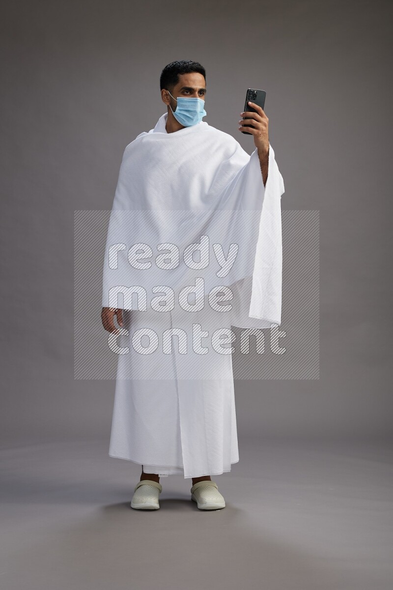 A man wearing Ehram with face mask Standing taking selfie on gray background