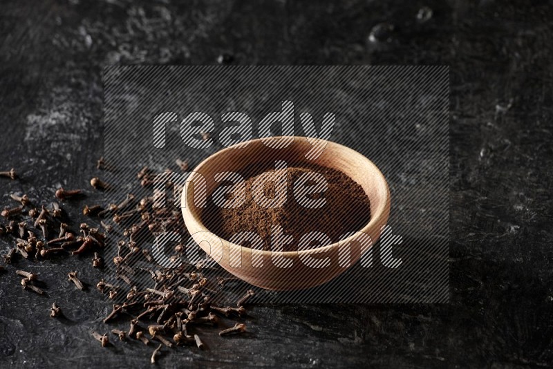 A wooden bowl full of cloves powder on a textured black flooring