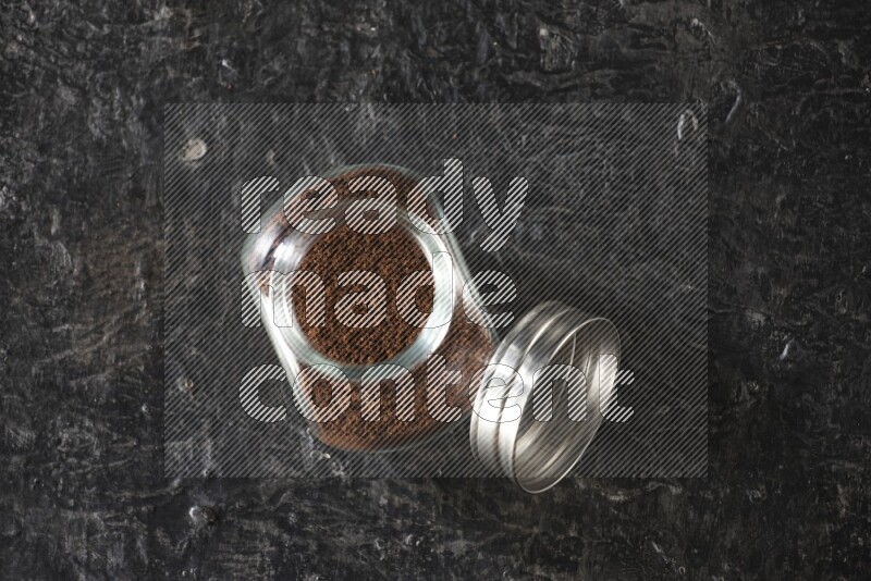 A glass spice jar full of cloves powder on textured black flooring