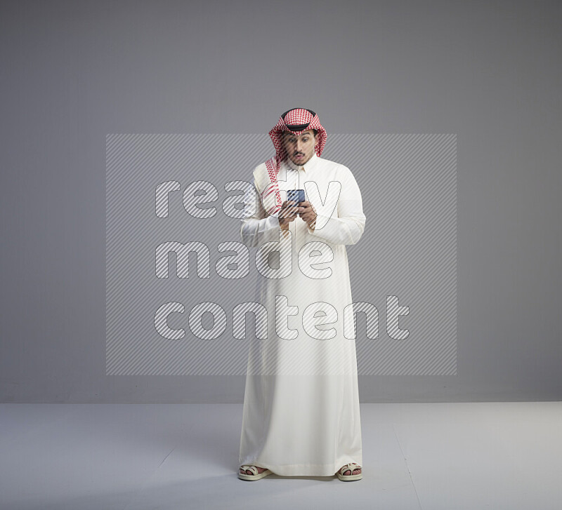 رجل سعودي يرتدي ثوب ابيض وشماغ احمر يمسك جواله بيده