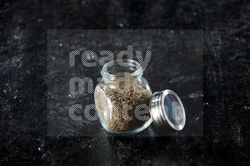 A glass spice jar full of cumin seeds on a textured black flooring