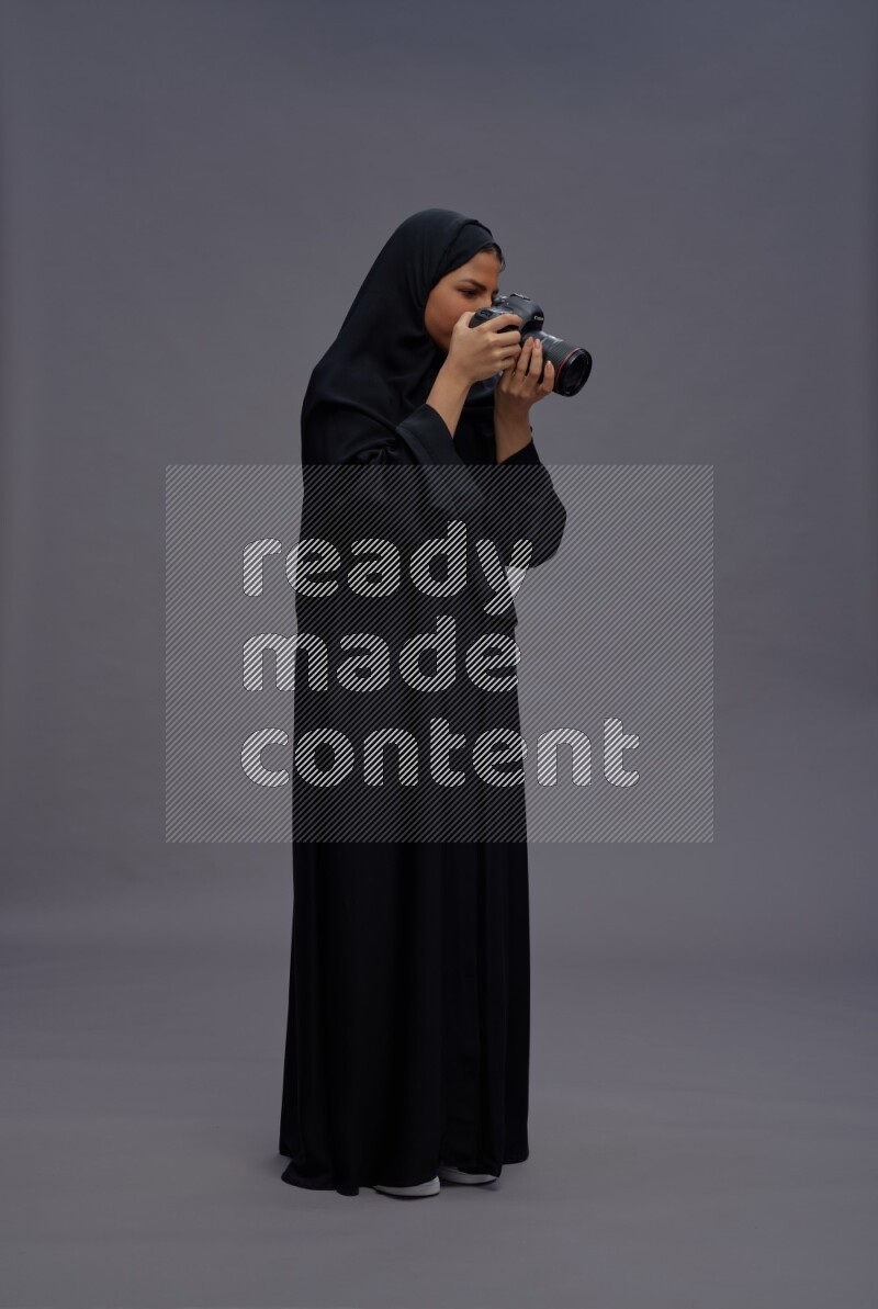 Saudi woman wearing Abaya standing holding Camera on gray background