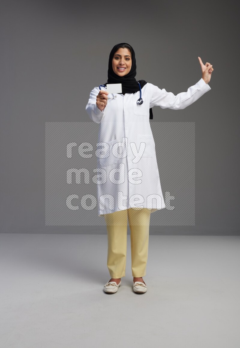 Saudi woman wearing lab coat with stethoscope standing holding ATM card on Gray background