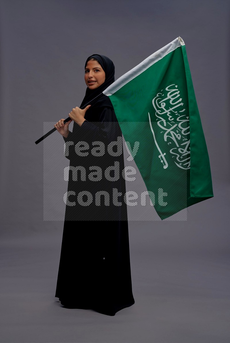 Saudi woman wearing Abaya standing holding Saudi flag on gray background