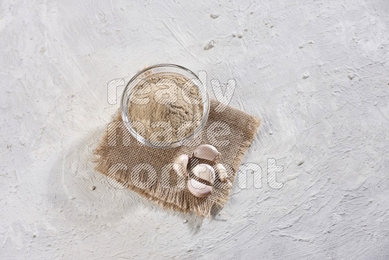 A glass bowl full of garlic powder on burlap fabric with garlic cloves on a textured white flooring in different angles