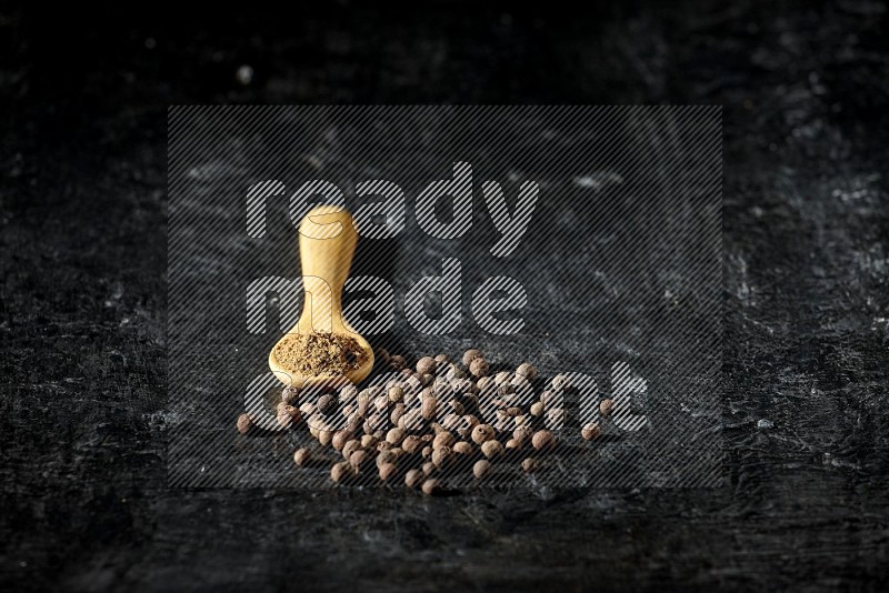 A wooden spoon full of allspice powder and whole balls spreaded on a textured black flooring