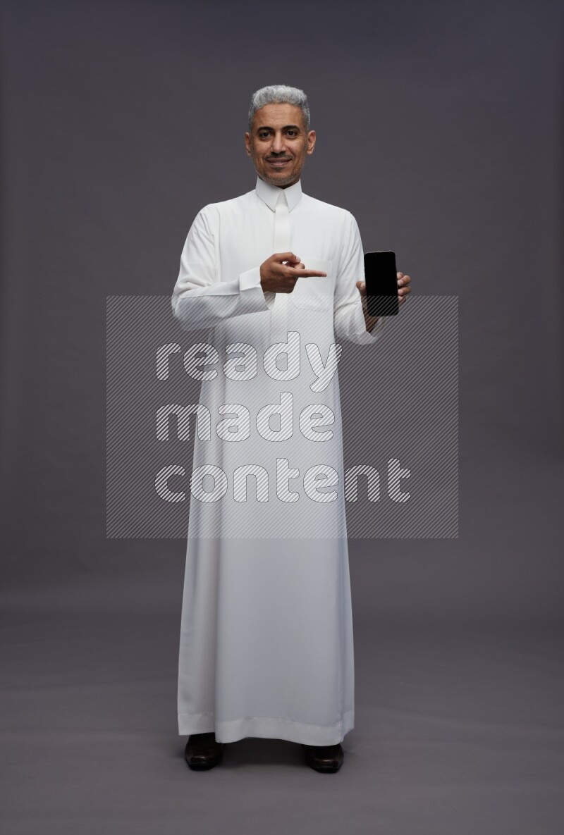 Saudi man wearing thob standing showing phone to camera on gray background