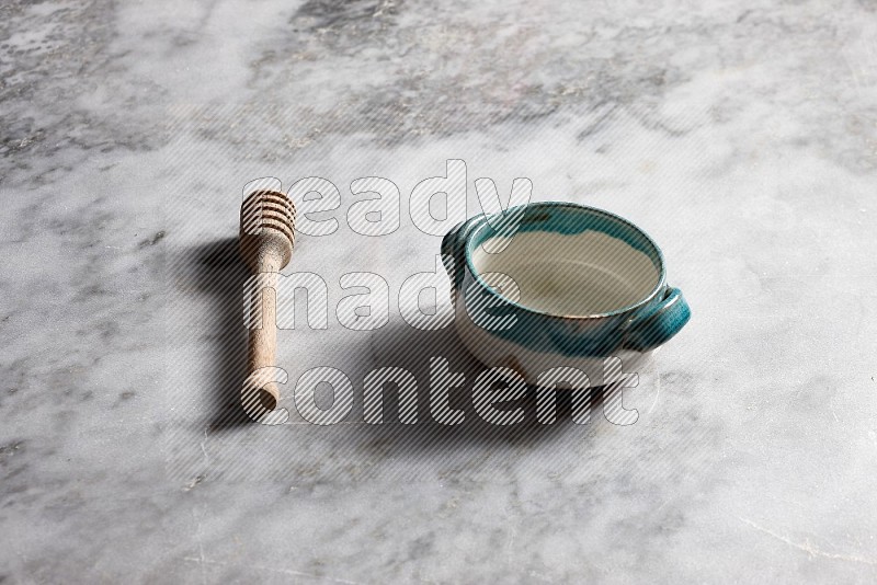 Multicolored Pottery bowl with wooden honey handle on the side with grey marble flooring, 45 degree angle