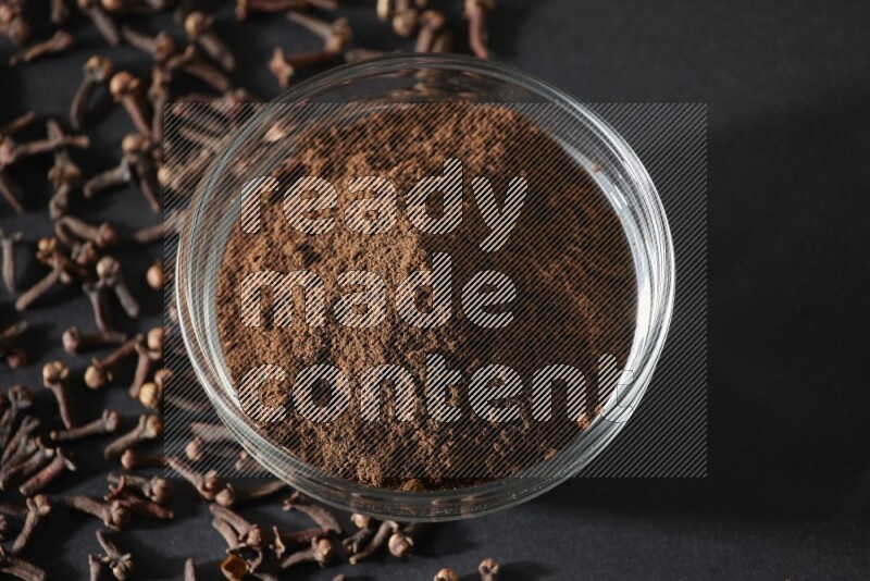 A glass bowl full of cloves powder with cloves grains spread on black flooring