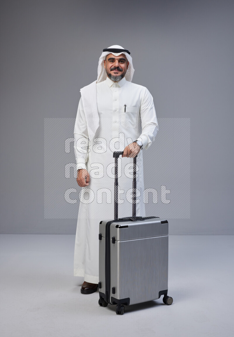 Saudi man wearing Thob and white Shomag standing holding Travel bag on Gray background