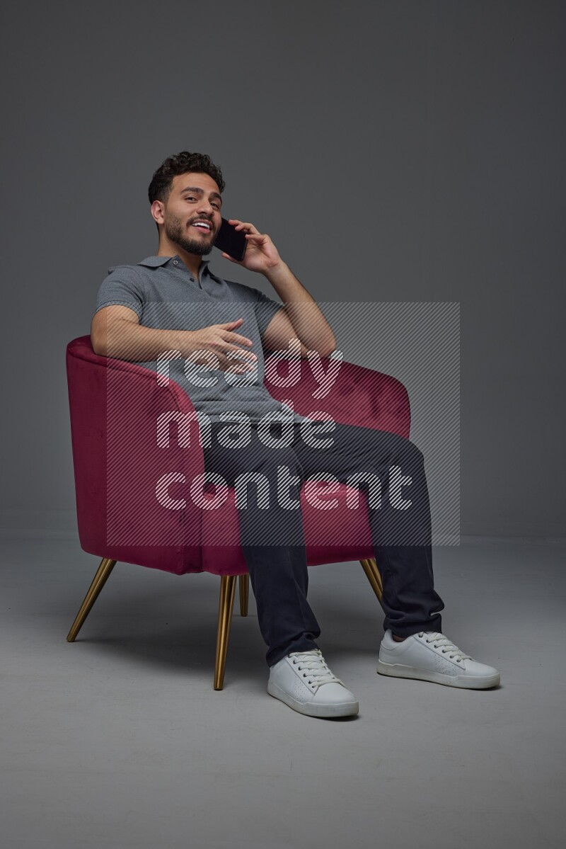 A man wearing casual and talking in his phone while setting on a burgundy chair eye level on a gray background