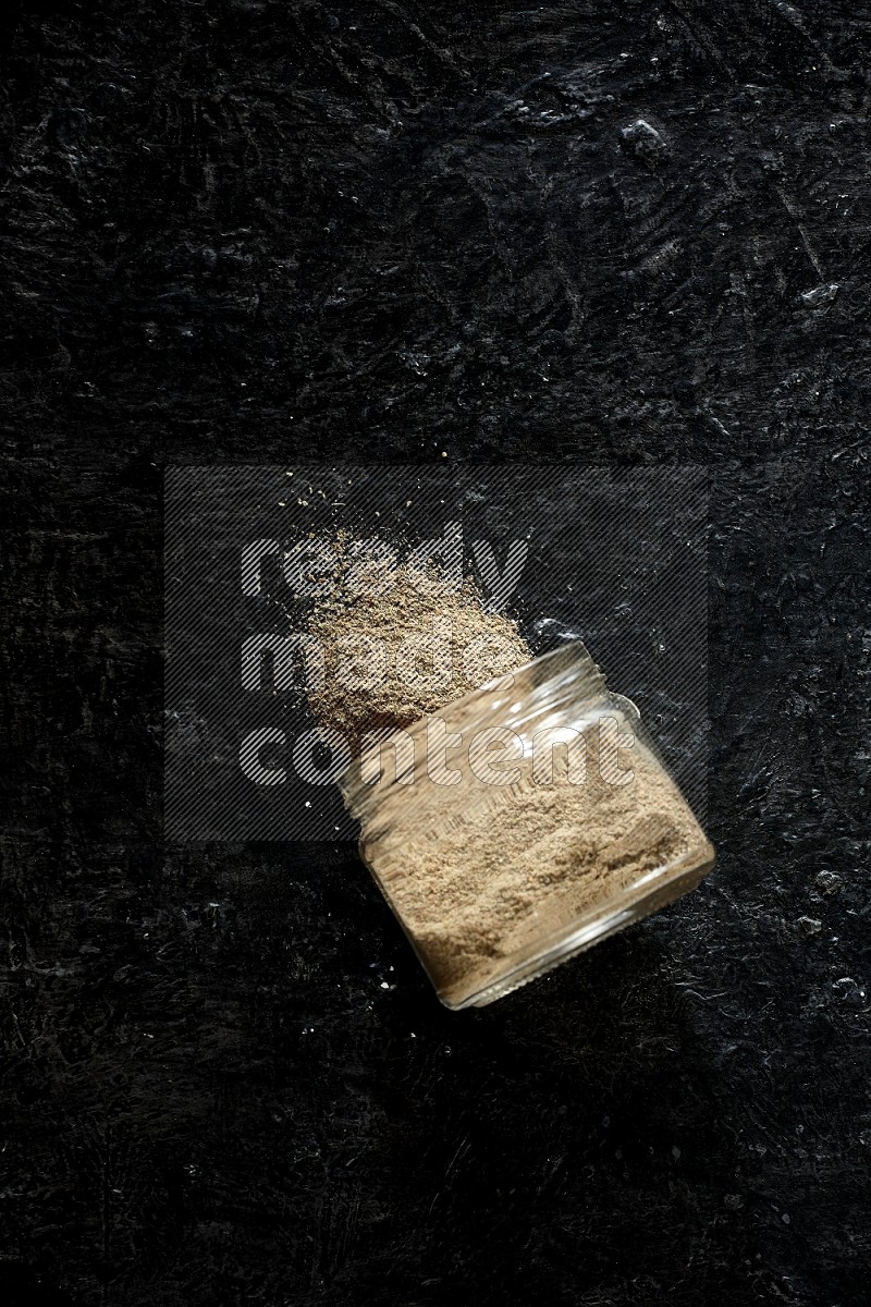 A flipped glass jar full of cardamom powder and powder spilled out of it on textured black flooring