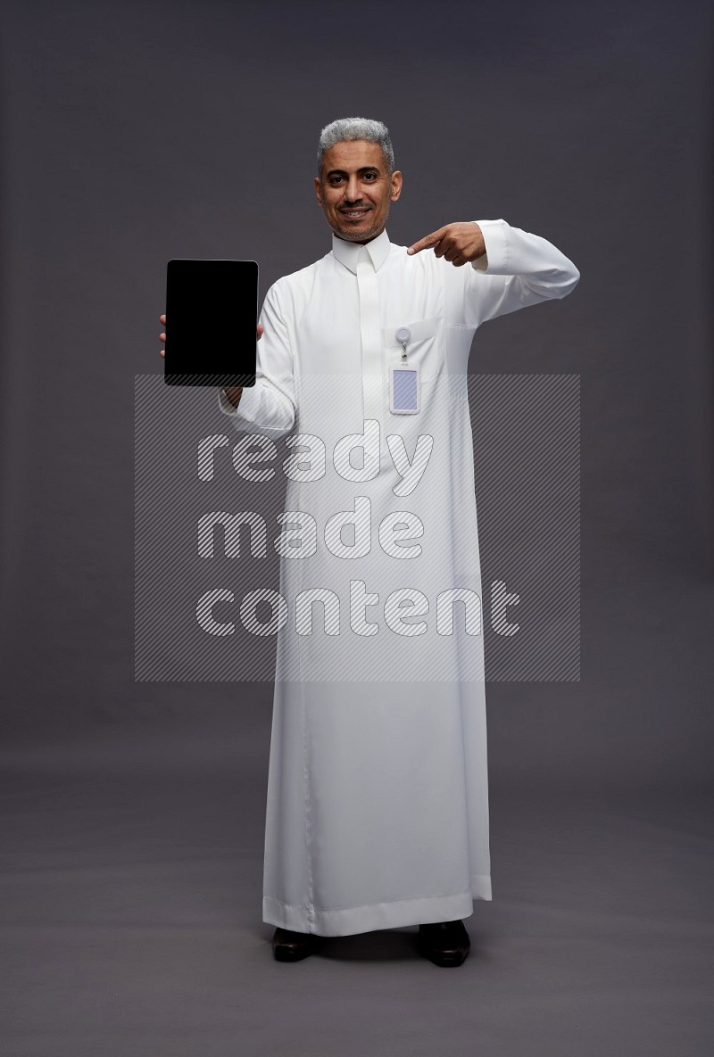 Saudi man wearing thob with pocket employee badge standing showing tablet to camera on gray background