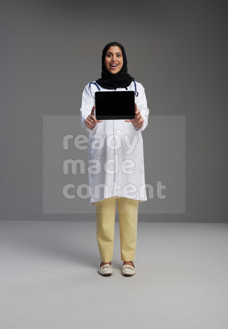 Saudi woman wearing lab coat with stethoscope standing showing tablet to camera with sign in the back on Gray background