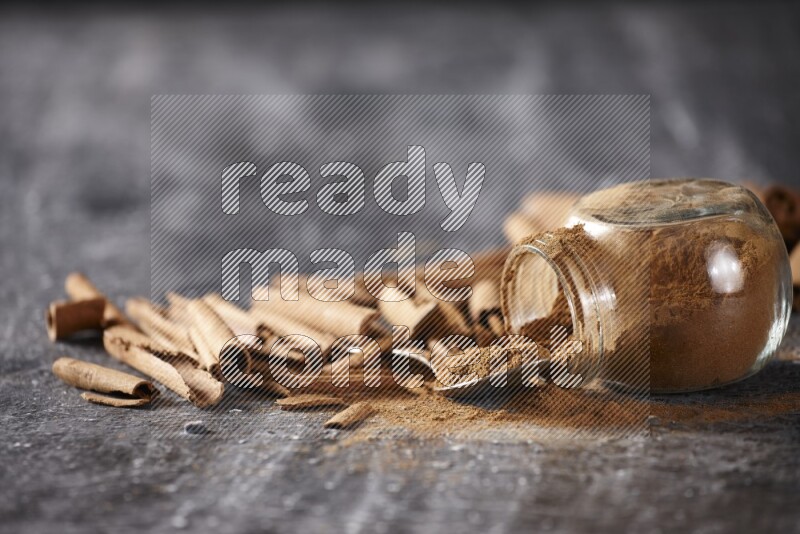Herbal glass jar full cinnamon powder flipped and a metal spoon full of powder surrounded by cinnamon sticks on textured black background in different angles