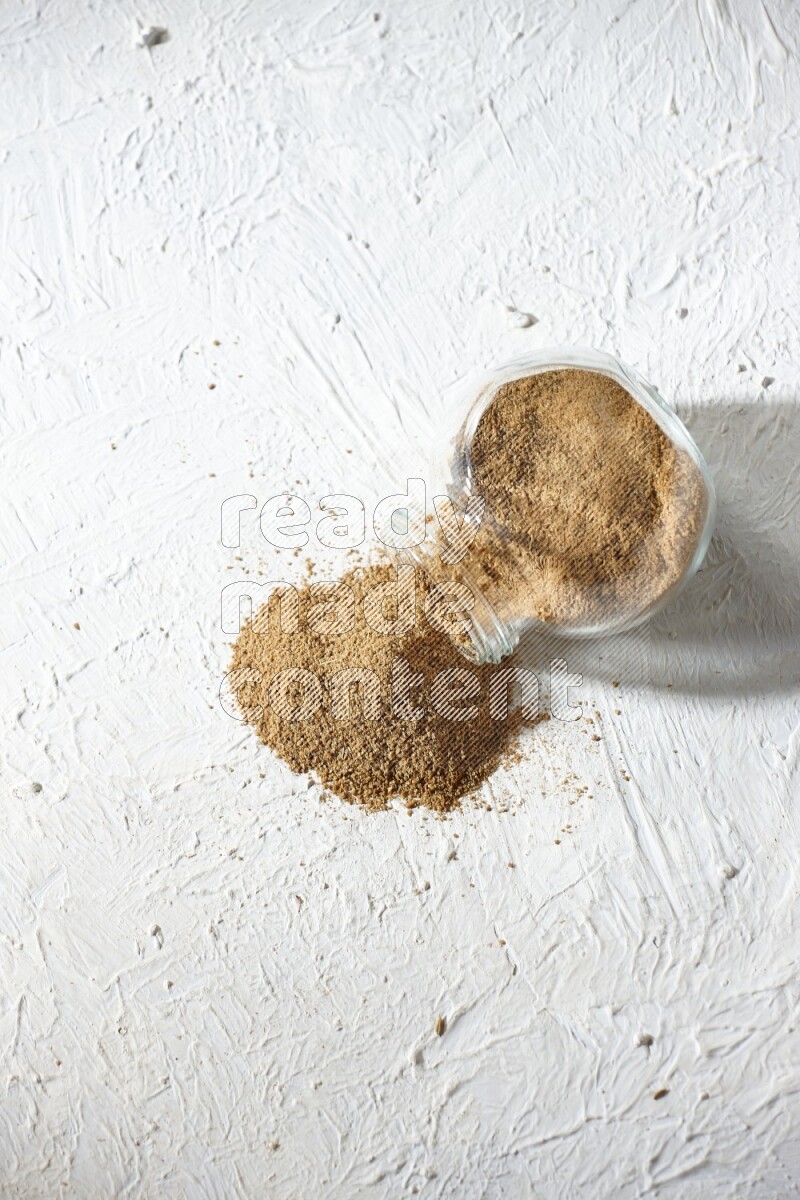 A flipped glass spice jar full of cumin powder and powder spilled out on textured white flooring
