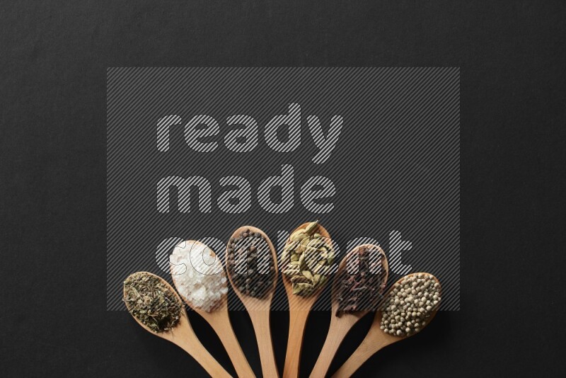 wooden spoons filled with white peppers, cloves, cardamom, salt, black peppers and basil on black flooring