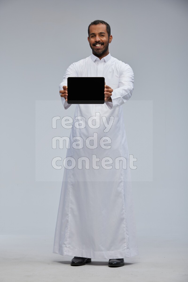 Saudi man wearing Thob standing showing tablet to camera on Gray background