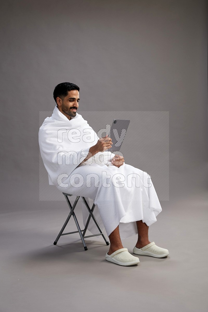 A man wearing Ehram sitting on chair holding a tablet on gray background