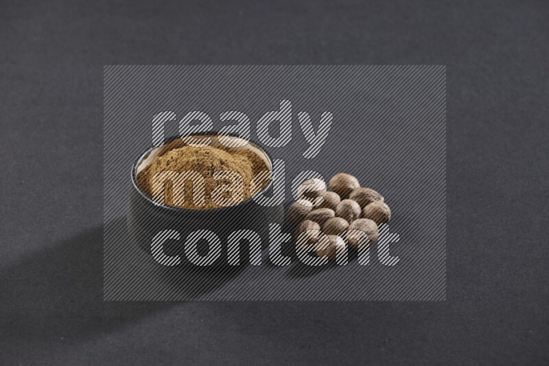 A black pottery bowl full of nutmeg powder with whole seeds beside it on a black flooring