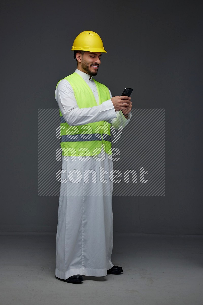 A Saudi man wearing Thobe with a yellow safety vest and white helmet standing and using his phone eye level on a gray background