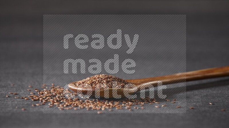 A wooden ladle full of flaxseeds and seeds spread beside it on a black flooring