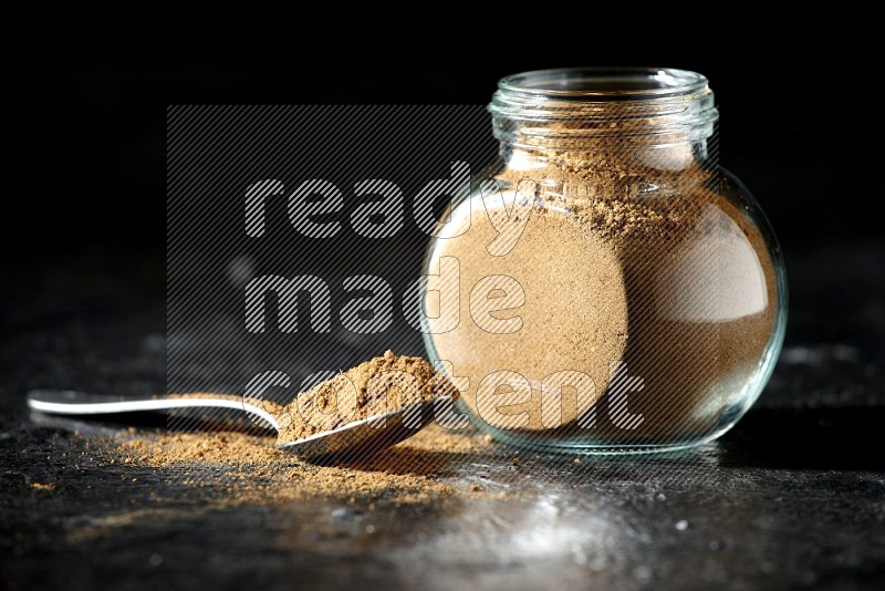 A glass spice jar and metal spoon full of allspice powder on a textured black flooring