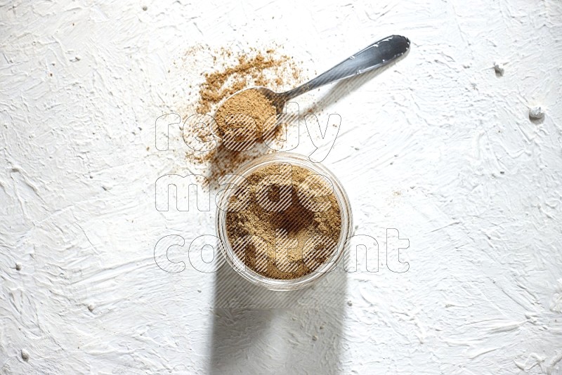 A glass jar and a metal spoon full of cumin powder on textured white flooring