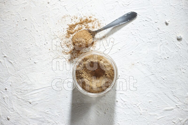A glass jar and a metal spoon full of cumin powder on textured white flooring