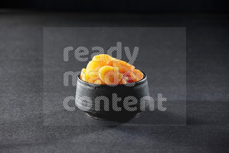 A black pottery bowl full of dried apricots on a black background in different angles