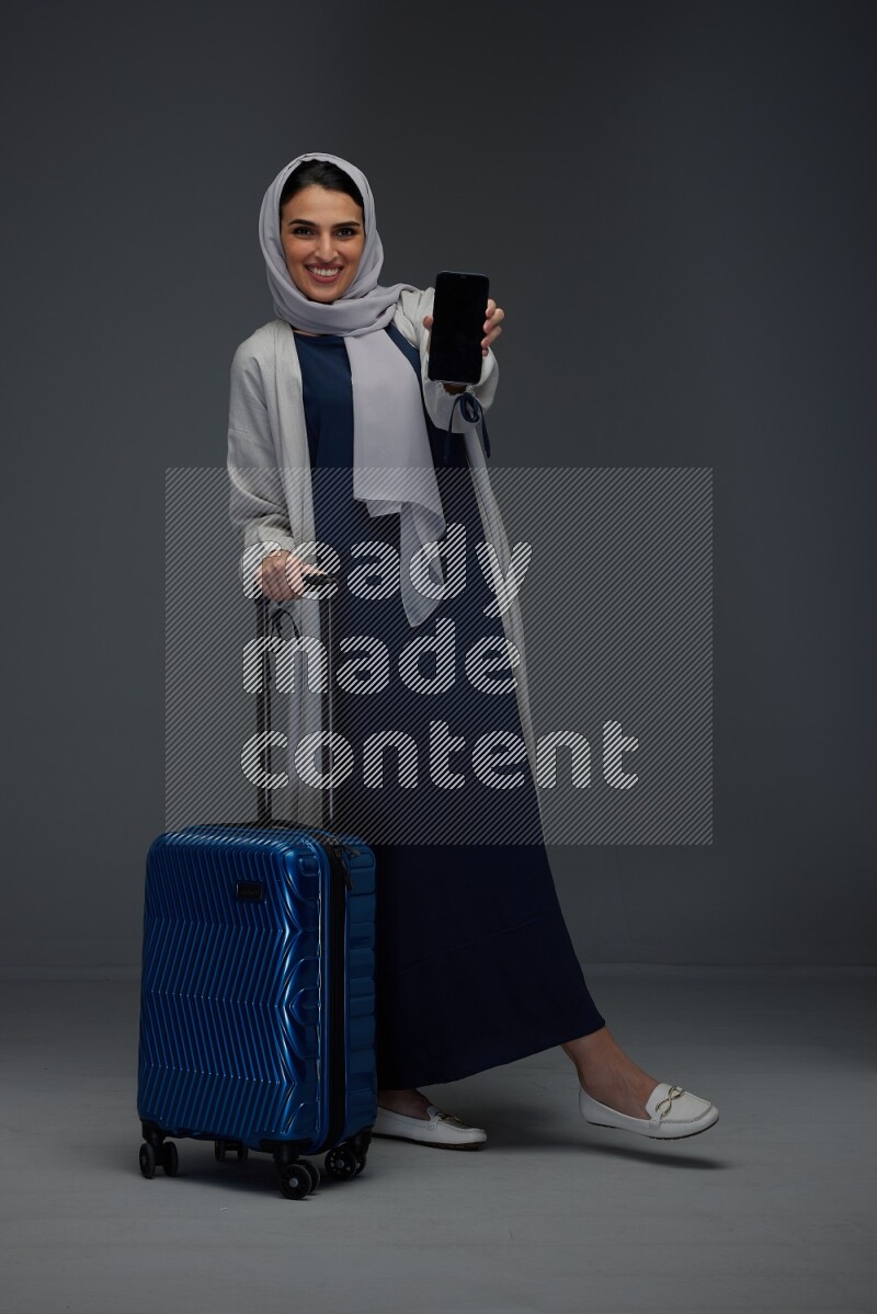 A Saudi woman wearing a light gray Abaya and head scarf standing and showing the phone's screen while wearing headphones eye level on a grey background