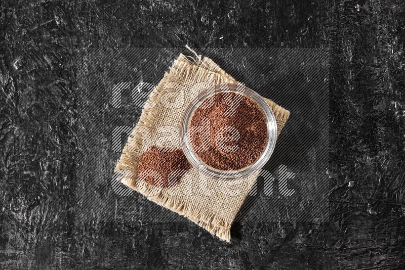 A glass bowl full of garden cress seeds with bunch of the seeds on burlap fabric on a textured black flooring