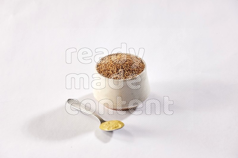 وعاء فخاري بيج ممتلئ ببذور الخردل وملعقة معدنية ممتلئة بمعجون الخردل علي خلفية بيضاء