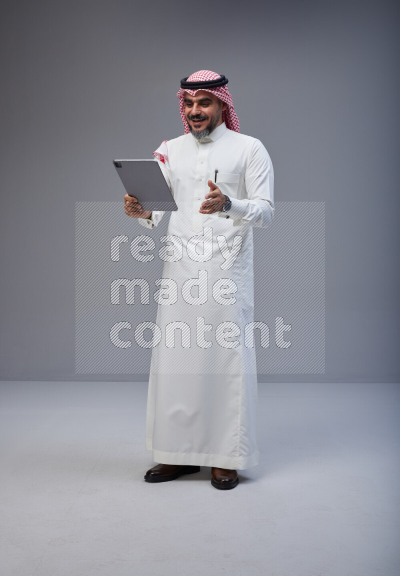 Saudi man Wearing Thob and red Shomag standing working on tablet on Gray background