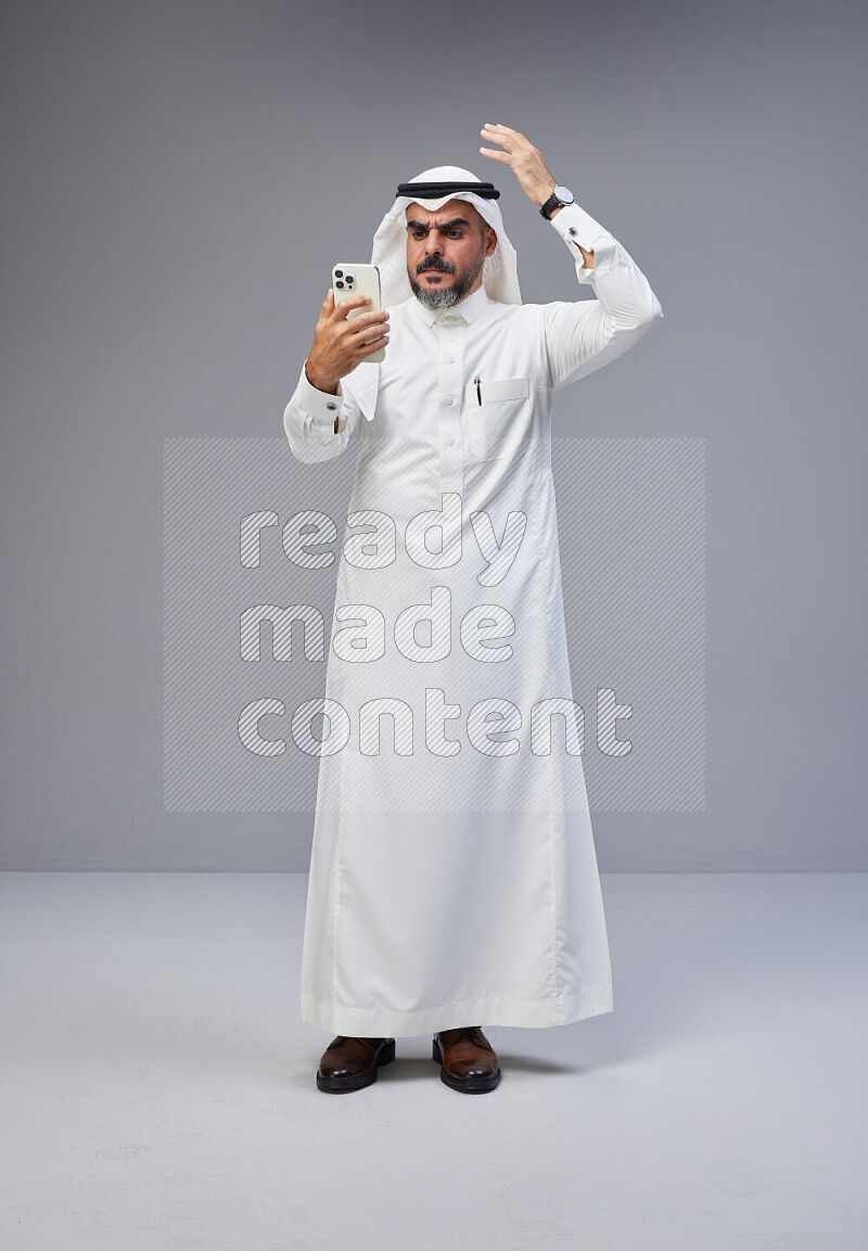 Saudi man Wearing Thob and white Shomag standing texting on phone on Gray background